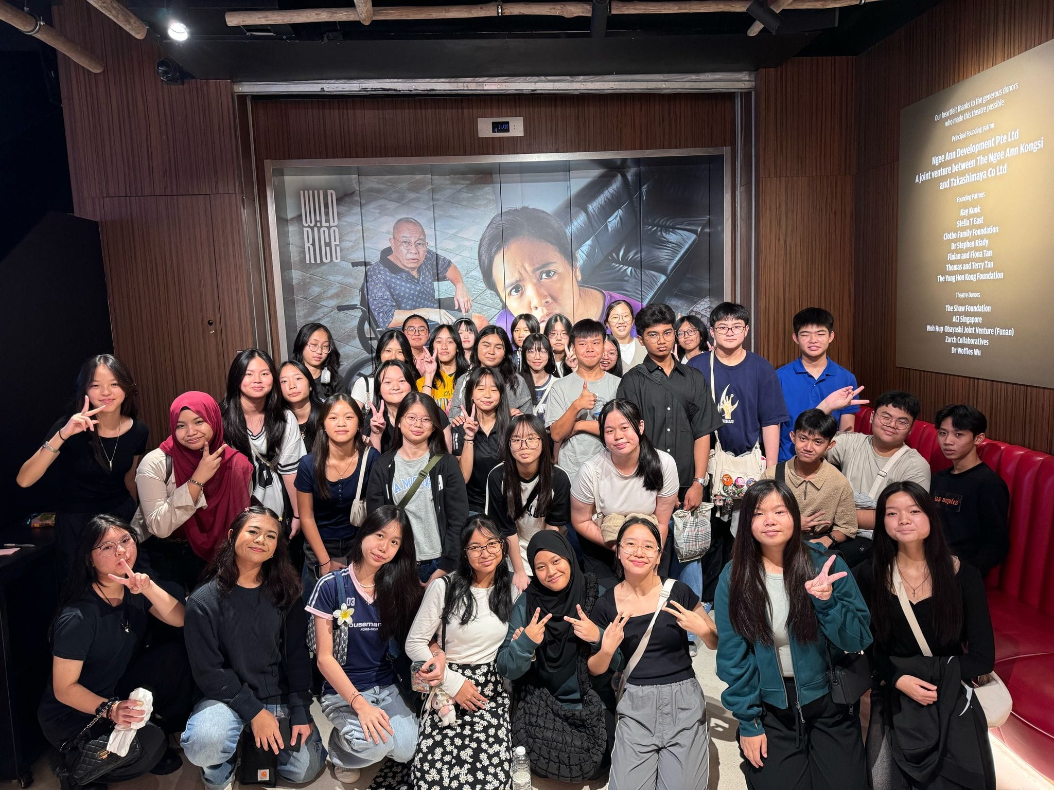 Secondary 3 students posing together for a group photo in a modern theatre lobby. In the background, there is a large "Wild Rice" poster featuring an older man in a wheelchair and a woman, alongside a donor appreciation plaque on a wooden wall.